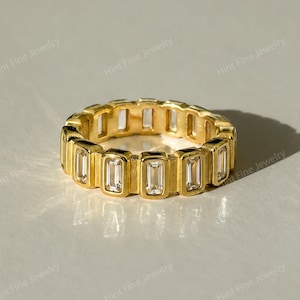 May include: A gold band ring featuring a continuous row of clear, rectangular baguette-cut stones set vertically within the metal frame. The ring displays the text Hint Fine Jewelry repeatedly across the image in a light, semi-transparent font.
