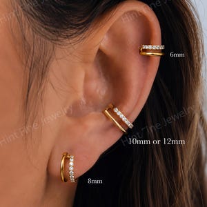 May include: Gold-coloured ear jewellery set. Includes a pair of 8mm hoop earrings with a row of small, clear stones, a 6mm ear cuff, and a 10mm or 12mm ear cuff, each with a row of clear stones. The jewellery is displayed on an ear.