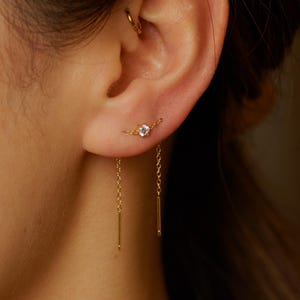 Puede incluir: Primer plano de un conjunto de pendientes dorados. El pendiente presenta una pequeña gema transparente conectada por una delicada cadena a una barra dorada colgante. El pendiente se lleva en la oreja, centrándose en la joya.