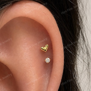 May include: Two gold-tone ear piercings are positioned on the upper cartilage. The top piece is a small, smooth, polished gold heart stud. The bottom piece is a small, round, clear, faceted gemstone stud set in a gold-tone metal prong setting.