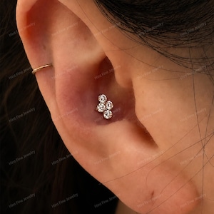 May include: A gold conch piercing stud featuring a cluster of four round, clear, brilliant-cut stones arranged in a floral-like pattern, positioned in the center of the ear, accompanied by a thin, plain gold hoop ring in the upper ear cartilage.