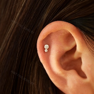 May include: Close-up of an ear with two small, round, diamond earrings. The earrings are set in gold and are positioned one above the other on the ear. The skin tone is light.