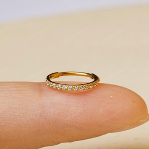 May include: A gold ring, adorned with a continuous line of small, clear gemstones. The ring is displayed on a finger, highlighting its intricate design and brilliance. This is a piece of jewellery.
