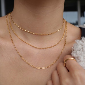 May include: Three gold chain necklaces layered on a person's neck. The chains are different styles and thicknesses. The top chain is a delicate, thin chain with small, oval-shaped links. The middle chain is a slightly thicker chain with a more traditional link style. The bottom chain is a thicker chain with larger, oval-shaped links.