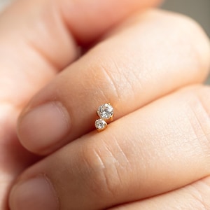 May include: A small, gold earring featuring two clear, round gemstones. The earring is held between fingers, highlighting its delicate design and sparkling stones. The jewellery piece is likely for a piercing.