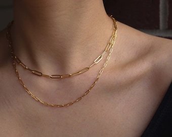 May include: Two gold chain necklaces layered on a person's neck. The top chain is a thicker, open link chain. The bottom chain is a thinner, closed link chain.