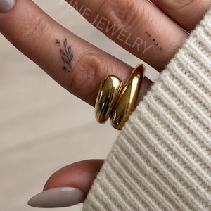 Peut inclure: Gros plan sur une main avec des ongles en amande peints dans une couleur taupe neutre. La main est ornée de deux bagues dorées et de petits tatouages d'un croissant de lune et d'une brindille de feuilles. Le texte "HINT FINE JEWELRY" est visible.