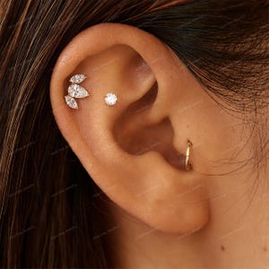 Peut inclure: Un ensemble de piercings d'oreille en or comprenant un groupe de trois pierres claires en forme de marquise et de poire disposées en feuille, un clou à pierre claire ronde et un anneau fin en or placé dans le cartilage inférieur.