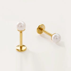 Peut inclure: Deux bijoux de piercing labret dorés avec une perle blanche. L'un est debout, montrant la base, la tige et la perle. L'autre est couché sur le côté, montrant la perle et la tige.