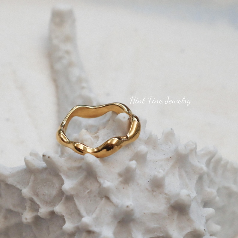 May include: A gold-toned ring with a wavy, organic design. The ring is displayed on a white, textured surface.