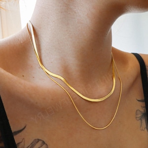 Peut inclure: Gros plan sur deux colliers dorés. Le collier supérieur est une chaîne serpent plate, et le collier inférieur est une chaîne fine et délicate. Les colliers sont portés sur le cou d'une personne, contre un haut noir.