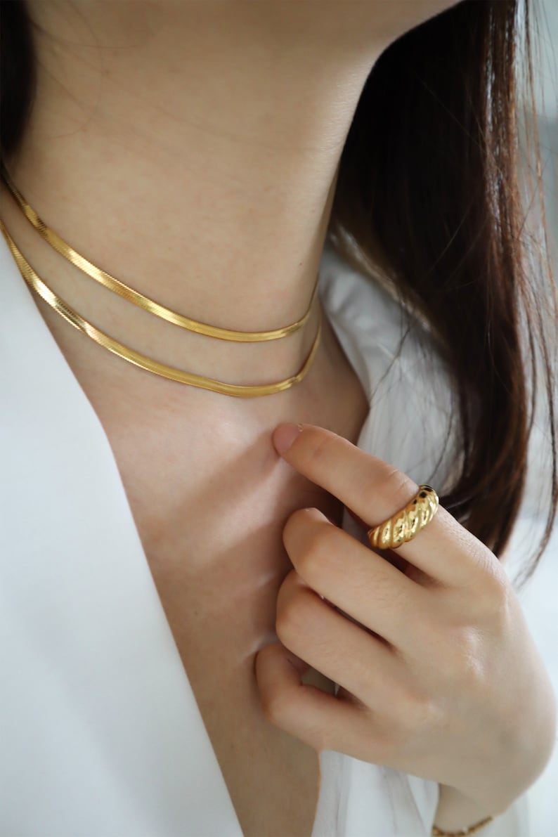May include: A close-up of a person wearing a gold chain necklace and a gold ring. The ring has a textured, rope-like design.