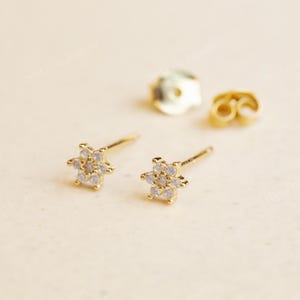 May include: Gold-coloured stud earrings, each with a flower design set with small, clear gemstones. The earrings are shown with their backing pieces on a light-coloured surface. The earrings are delicate and elegant.