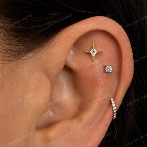 May include: A gold-toned ear piercing set featuring a four-pointed star stud with a central clear stone, a small round clear stone stud, and a silver-toned hoop earring lined with small clear stones, all worn in a single ear cartilage.