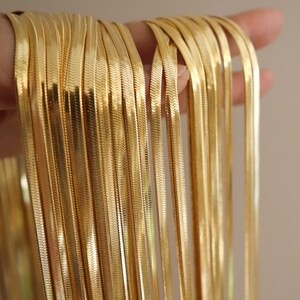 May include: A close-up of a stack of gold-colored snake chain necklaces. The chains are all the same length and width, and they are arranged in a neat pile.