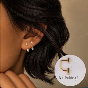 May include: A close-up of an ear with three pairs of gold earrings with clear, sparkling stones. The earrings are in various shapes, including round and marquise. A gold earring with a clear stone is shown with the text "No Poking!"