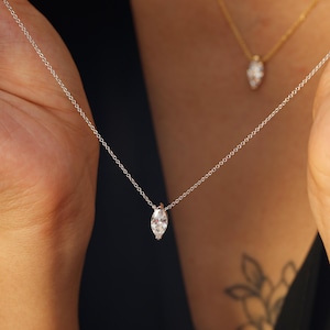 May include: A silver necklace with a marquise-cut diamond pendant. The delicate chain is held up by a person. A second gold necklace with a similar pendant is visible in the background. The jewelry is set against a dark background.