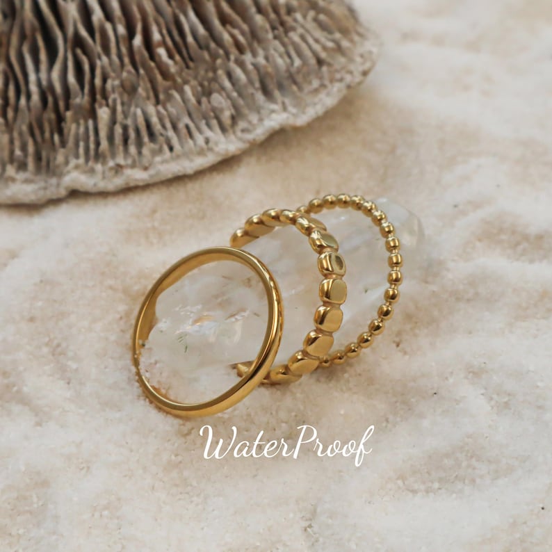 K&ouml;nnte beinhalten: Ein Set aus drei goldfarbenen Ringen, die auf einem klaren Kristall liegen. Ein Ring hat eine glatte Schiene, einer eine texturierte Oberfl&auml;che mit kleinen abgerundeten Formen und einer ist ein zartes Perlenband. Der Text "WaterProof" ist in Wei&szlig; gedruckt.