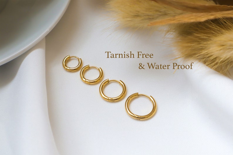May include: Four gold hoop earrings of different sizes on a white fabric background. The text "Tarnish Free & Water Proof" is written in black text on the background.