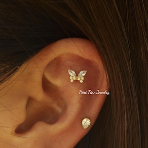 May include: An ear adorned with two pieces of jewellery. A butterfly-shaped stud with clear stones and gold metal is visible. A teardrop-shaped stud with a clear stone is also present. The text "Hint Fine Jewelry" is displayed.