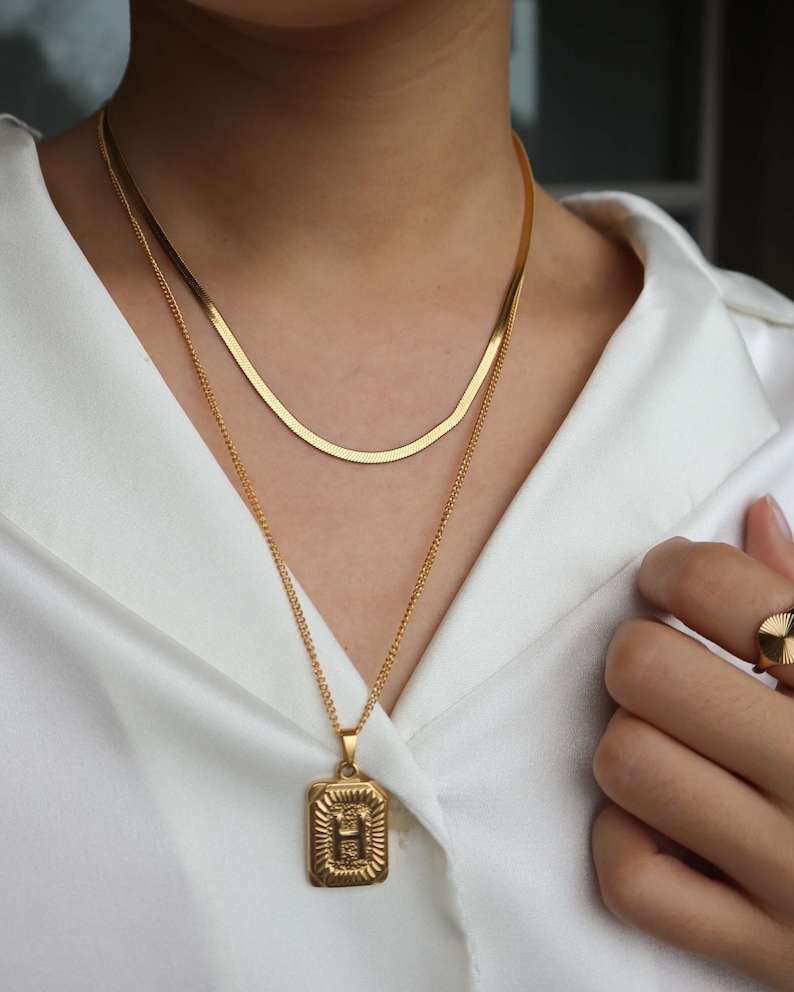 May include: Two gold necklaces, one with a thin chain and a rectangular pendant with the letter "H" on it, and the other with a thicker, flat chain. The necklaces are layered on a person's neck.