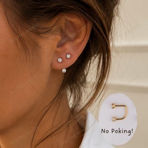 May include: An ear with three small, round, clear stud earrings. A gold-coloured earring with a clear stone at each end is shown in a circular graphic with the text "No Poking!"