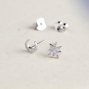 May include: A pair of silver stud earrings. One earring is a crescent moon shape, and the other is a starburst design. Both earrings are embellished with small, clear gemstones. Earring backs are also visible.