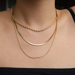 May include: Three gold chains layered on a person's neck. The chains are different styles, including a rope chain and a snake chain.