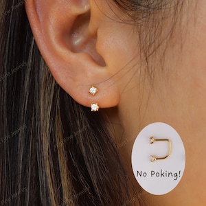 May include: A close-up of a person's ear with two small, gold-colored stud earrings, each featuring a clear, round gemstone. The earrings are set close to the ear. A small graphic with the text "No Poking!" is in the lower right corner.