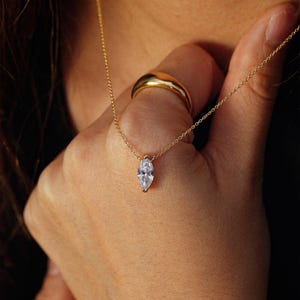 May include: A delicate gold necklace with a marquise-cut clear gemstone pendant. A gold ring is on the finger. The jewelry is set against a warm, neutral background, highlighting the sparkle of the pendant and the shine of the gold.