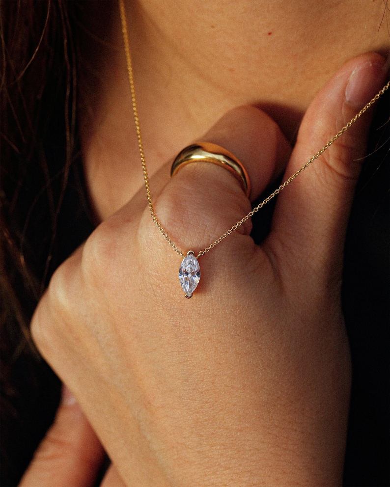 May include: A delicate gold necklace with a marquise-cut clear gemstone pendant. A gold ring is on the finger. The jewelry is set against a warm, neutral background, highlighting the sparkle of the pendant and the shine of the gold.