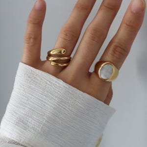 May include: Two gold rings on a hand. One ring is a simple band with a white oval stone. The other ring is a twisted band.