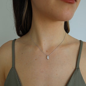 May include: A silver necklace with a small, pear-shaped pendant. The pendant is set with a clear, sparkling stone. The necklace is worn on a person's neck, against a green tank top.