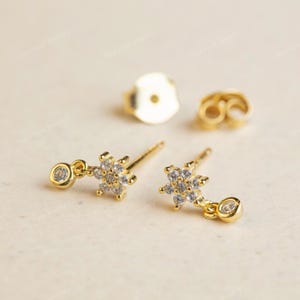 May include: Gold-tone stud earrings with a snowflake design. Each earring has a small, round, clear gemstone at the end of a fine chain, and a larger snowflake shape with multiple clear gemstones. The earrings are presented on a light beige surface.