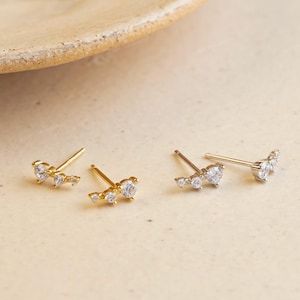 May include: Gold and silver stud earrings featuring clear cubic zirconia stones. The earrings are arranged in a curved line, with a larger stone at the center. The earrings are displayed on a neutral surface. The earrings are made of gold and silver.