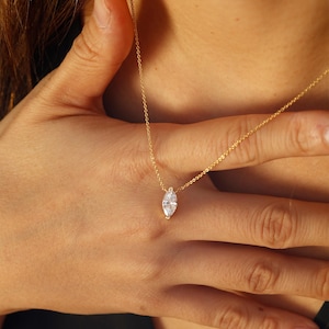 May include: A delicate gold necklace featuring a marquise-cut gemstone pendant. The pendant is a clear, sparkling stone set in a gold bezel. The necklace is shown against a skin tone background.