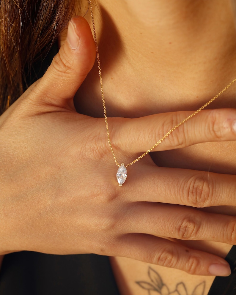 May include: A delicate gold necklace featuring a marquise-cut gemstone pendant. The pendant is a clear, sparkling stone set in a gold bezel. The necklace is shown against a skin tone background.