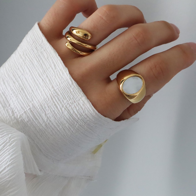 May include: Two gold rings on a hand. One ring is a thick band with a white oval stone. The other ring is a thin, twisted band.