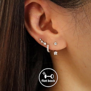 May include: An ear displaying several silver earrings. There's a curved bar of three small, round, clear earrings, a small hoop earring with clear stones, a single round clear stud, and a square clear stud. The image also shows a 'flat back' logo.