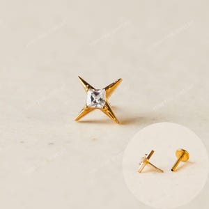 May include: A gold stud earring featuring a four-pointed star design with a central square-cut clear gemstone. The inset shows the earring from the side, revealing the post and a flat circular backing piece for secure attachment.