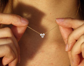 May include: A delicate gold necklace featuring three small, clear gemstones arranged in a cluster. The necklace is held up against a person's skin, showcasing the jewelry's design and the chain's fine details. The necklace is a piece of jewelry.