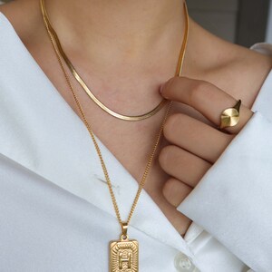 May include: A gold chain necklace with a pendant featuring the letter "H" in a square frame. The necklace is layered over a thinner gold chain. A gold ring with a round face is also visible.
