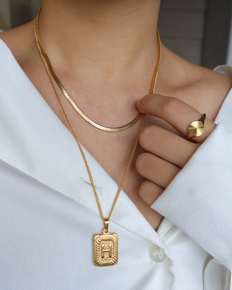 May include: A gold chain necklace with a pendant featuring the letter "H" in a square frame. The necklace is layered over a thinner gold chain. A gold ring with a round face is also visible.