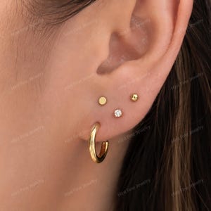 Peut inclure: Un ensemble de quatre boucles d'oreilles dorées comprenant une petite créole, un clou plat rond, un petit clou sphérique et un clou rond orné d'une pierre précieuse blanche transparente au centre du lobe de l'oreille.