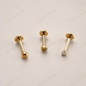 May include: Three gold-toned flat back labret studs. The first stud features a smooth spherical end, the second is a plain flat disc end, and the third is topped with a small, clear, square-cut gemstone. Each piece has a thin post.