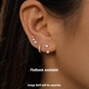 May include: A close-up of an ear adorned with multiple pieces of gold jewelry. The ear features a graduated stud earring, a small hoop earring, and two pairs of stud earrings, all with clear, sparkling stones. The text "Flatback available" is visible.