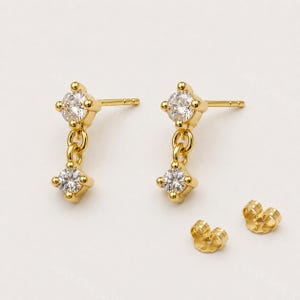 May include: A pair of gold stud earrings with a dangling design. Each earring has a clear, square gemstone at the top and bottom, linked by a small chain. Two additional earring backs are also visible.