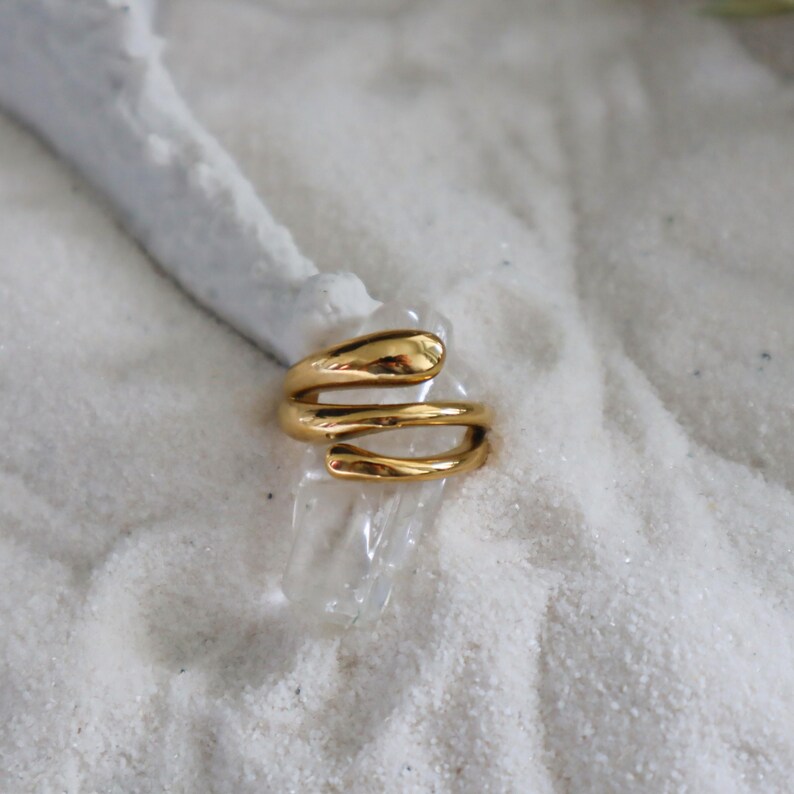 May include: A gold-toned ring with a unique, twisted design. The ring is made of a smooth, polished metal and has a modern, minimalist aesthetic.