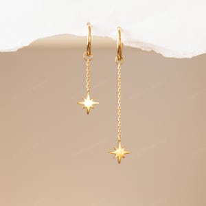 May include: A pair of gold-coloured hoop earrings. Each earring has a delicate chain suspending a star-shaped charm. The earrings are displayed against a neutral background, highlighting their elegant design. The earrings are a stylish accessory.