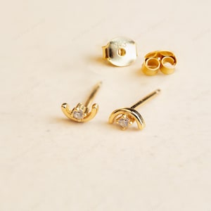 May include: Gold-coloured crescent moon stud earrings, each with a small, clear gemstone. The earrings are shown with their backing pieces on a light surface. These earrings are a delicate piece of jewellery.
